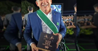 Governador Mauro Mendes sorrindo e segurando a Comenda da Ordem do Mérito Anhanguera, com uma faixa verde e branca no peito, em evento noturno.