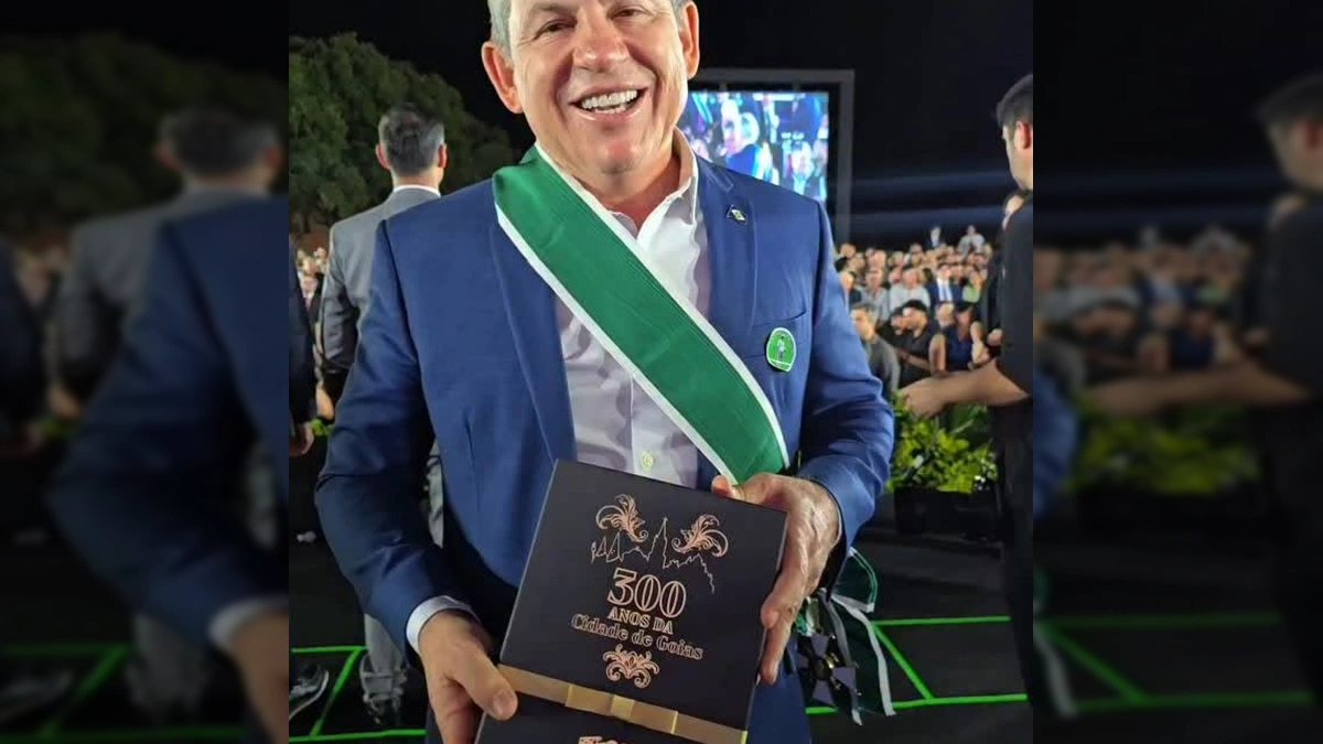 Governador Mauro Mendes sorrindo e segurando a Comenda da Ordem do Mérito Anhanguera, com uma faixa verde e branca no peito, em evento noturno.