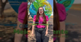 Mulher de óculos e camisa rosa vibrante falando sobre o evento ambiental em Tangará da Serra, com logo da corrida em destaque