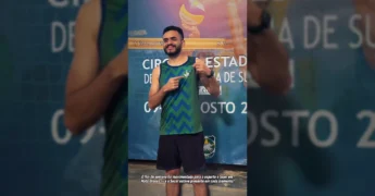 Homem sorrindo e apontando para a camisa com o logo da Federação Mato-grossense de Desportos de Surdos, em evento de vôlei de praia para surdos, com créditos para SecelMT no Instagram