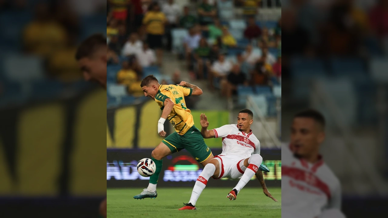 Imagem de um jogador do Cuiabá, camisa amarela e verde, em lance de ataque contra um jogador do CRB, camisa branca, durante partida da Série B. Crédito: AssCom Dourado / Instagram fmfmt_oficial